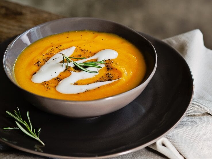 Dunkler Teller mit Kürbissuppe, garniert mit Kokosmilch und Rosmarin. 