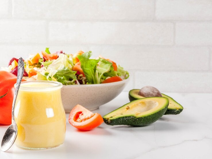 Ein Glas mit Honig-Senf-Dressing steht auf einem weißen Tisch, dahinter eine schale Salat.  | © iStock/Rimma_Bondarenko