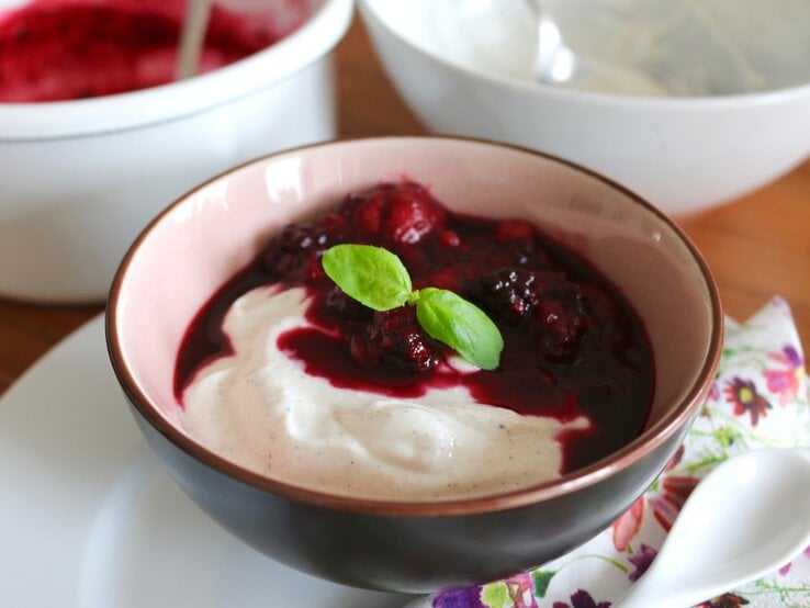 Quark-Dessert mit Vanille in einer dunklen Schüssel auf einem weißen Unterteller, rechts davon ein weißer Löffel und der Hintergrund ist hölzern.