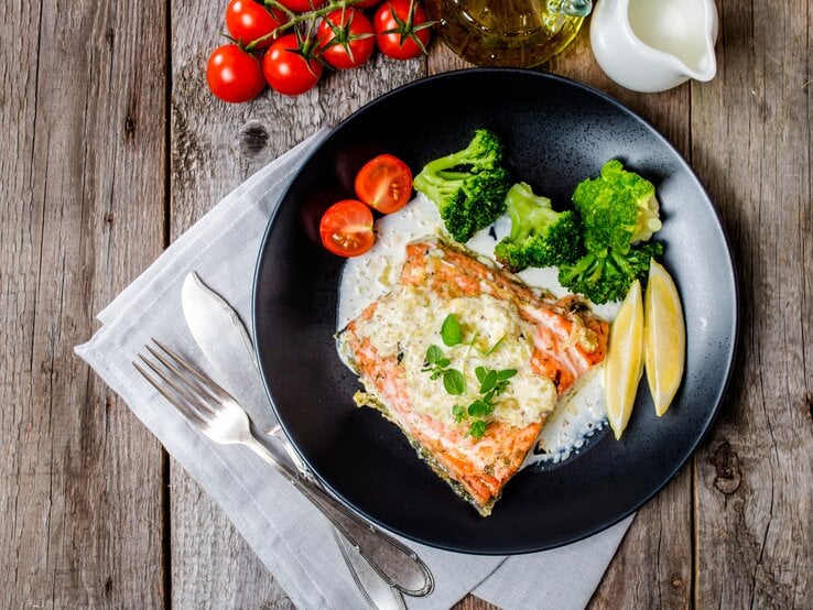 Draufsicht auf einen schwarzen Teller mit einem Stück Lachs und einer cremigen Weißweinsoße. Daneben Zitronenschnitze, Tomaten und Brokkoli. Das ganze steht auf einem dekorierten Holztisch.  | © iStock/ Roxiller
