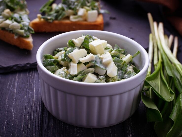 Bärlauch-Salat mit Käse in einer weißen Schale, angerichtet auf einem dunklen Holztisch. 