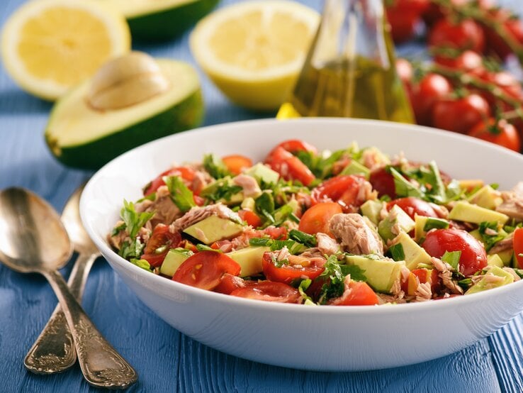 Weißer Teller mit Avocado-Thunfisch-Salat, Tomaten und Petersilie auf blauem Untergrund – umrundet von den Zutaten für den Salat.