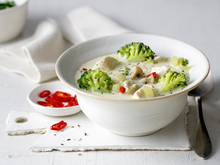 Eine weiße Schale mit der Hähnchen-Brokkoli-Suppe auf einem weißen Tisch von der Seite fotografiert.