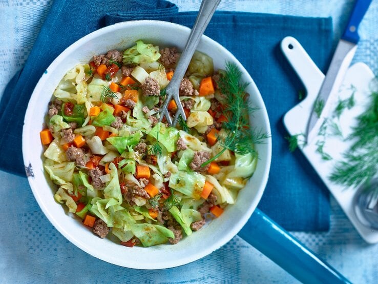 Spitzkohl-Hack-Pfanne mit Stil / Pfannenwender / Daneben Brettchen, Küchenmesser und frische Kräuter / Draufsicht