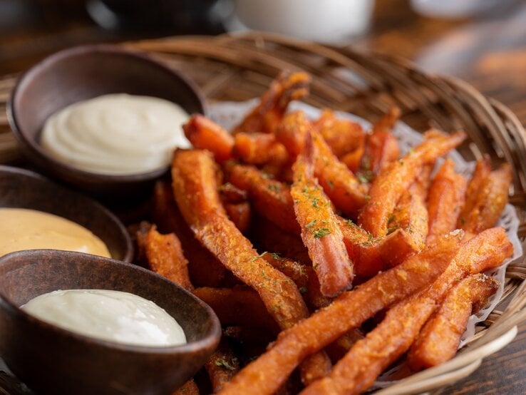 Süßkartoffelpommes aus der Heißluftfritteuse in Bambusschale mit dreierlei Dips in braunen Schalen auf Holztisch. Draufsicht.