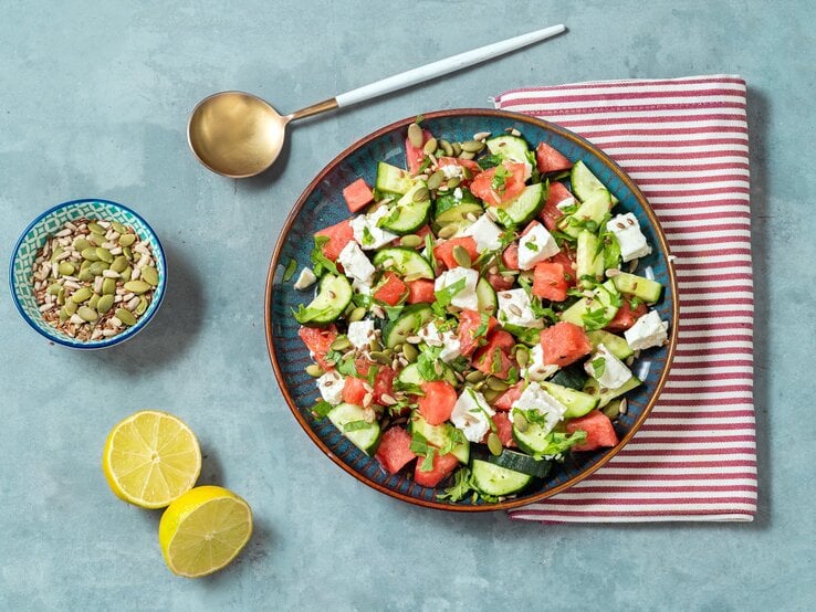 Gurkensalat mit Wassermelone und Feta auf blauem Teller, rot-weißem Tuch und hellblauem Untergrund. Im BIld eine Lössel, eine Schale mit Kernen und eine geteilte Zitrone. Draufsicht. | © Getty Images / Carlo A