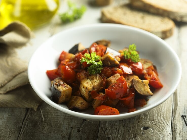 Italienischer Auberginen-Eintopf Caponata in weißem Teller auf rustikaler Oberfläche.