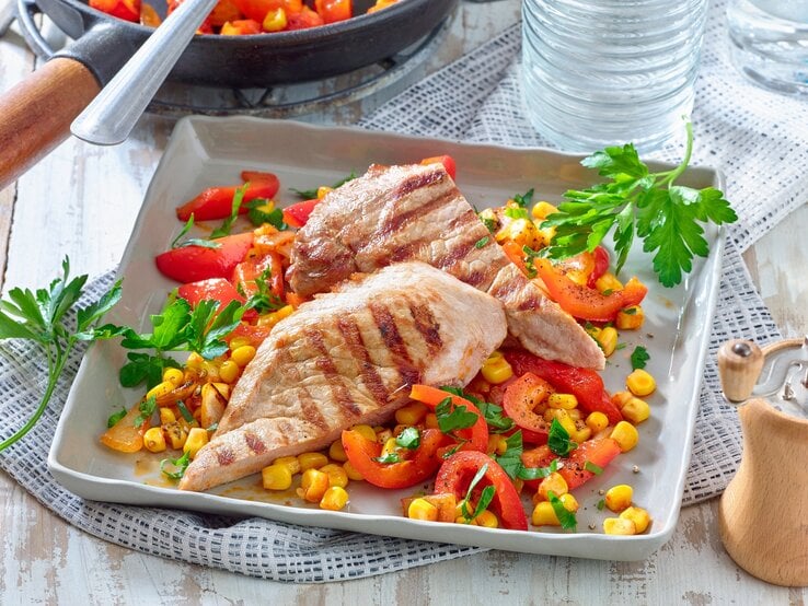 Schnitzel mit Mais und Paprika und Petersilie / quaratischer Teller / Tischdecke / Pfeffermühle aus Holz / Draufsicht schräg