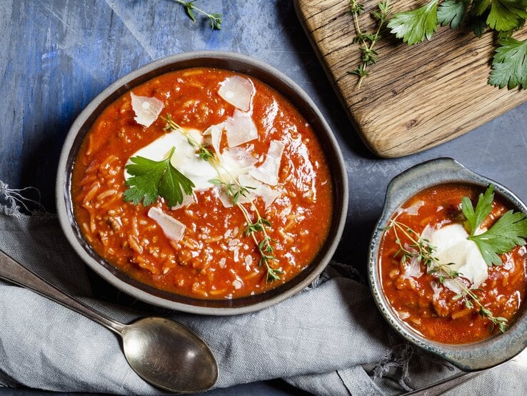 Tomatensuppe mit Reis, Hackfleisch und Parmesanspänen in einer grauen Schale auf einem dunkelblauen Steintisch. Alles direkt von oben fotografiert.