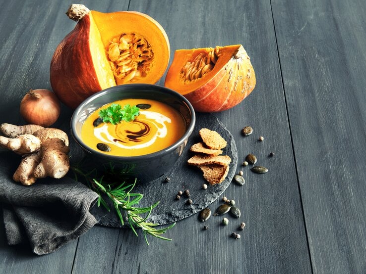 Schwarze Schale gefüllt mit Kürbsisuppe, garniert mit Creme und Kürbiskernen. Daneben liegt ein angeschnittener Kürbis. 