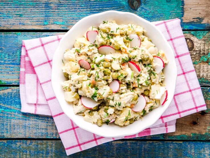 Falscher Kartoffelsalat mit Radieschen und Kräutern in weißer Schale auf rot-weißer Serviette, rustikaler Holztisch. | © iStock/wmaster890