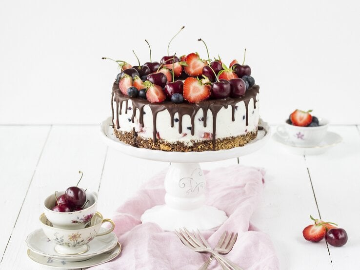 Weiße Beerentorte mit Kirschen und Erdbeeren auf einem Tortenständer, mit Schokoladenglasur am Rand. Die Torte steht auf einem hellen Holztisch, umgeben von Vintage-Tassen, Besteck und einem rosa Tuch.