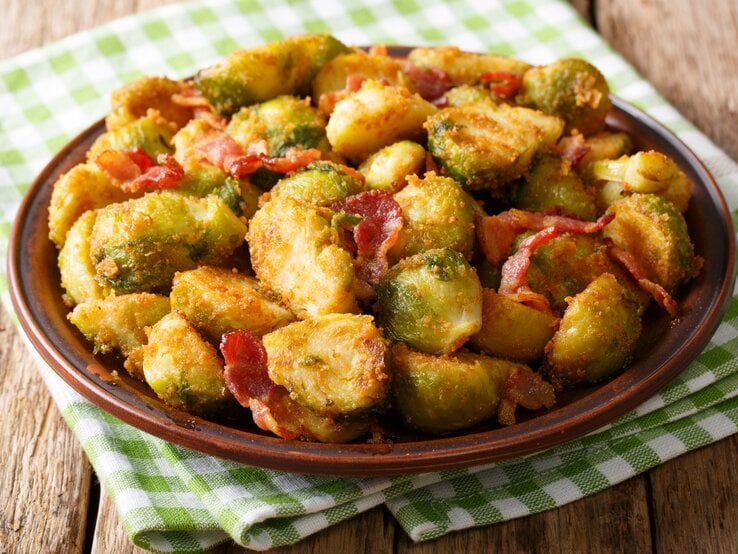 Brauner Teller belegt mit gebratenem Rosenkohl, Speck und Semmelbröseln. Das Ganze steht auf einem Holztisch.  | © iStock/ALLEKO