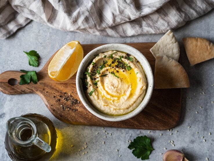 Mangold-Hummus: eine Schale mit Hummus, Olivenöl und Petersilie auf einem Brett, daneben ein Stück Zitrone, einige Scheiben Pitabrot, ein Kännchen mit Öl und ein Handtuch. 
