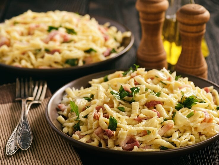 Draufsicht, schräg: Zwei runde Teller auf dem längliche Nudeln, kurze Speckstreifen und frische Petersilie liegen. Daneben liegen zwei Gabeln. Im Hintergrund zwei zwei Gewürzmühlen aus Holz.