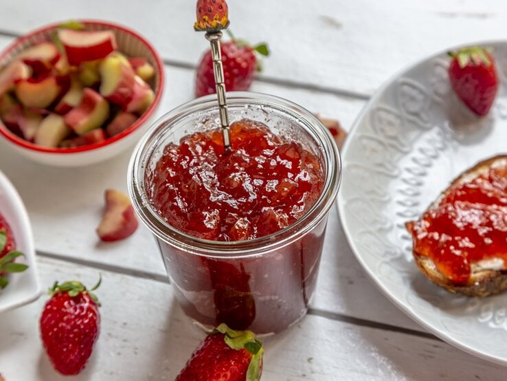 Draufsicht, schräg: Ein Glas Rhabarber-Erdbeer-Gelee steht auf einem weißen Untergrund aus Holz. Daneben liegen Erdbeeren, Rhabarerstücke sowie ein weißer Teller mit einem weißen Teller, auf dem eine Scheibe Brot mit Rhabarber-Erdbeer-Gelee liegt.