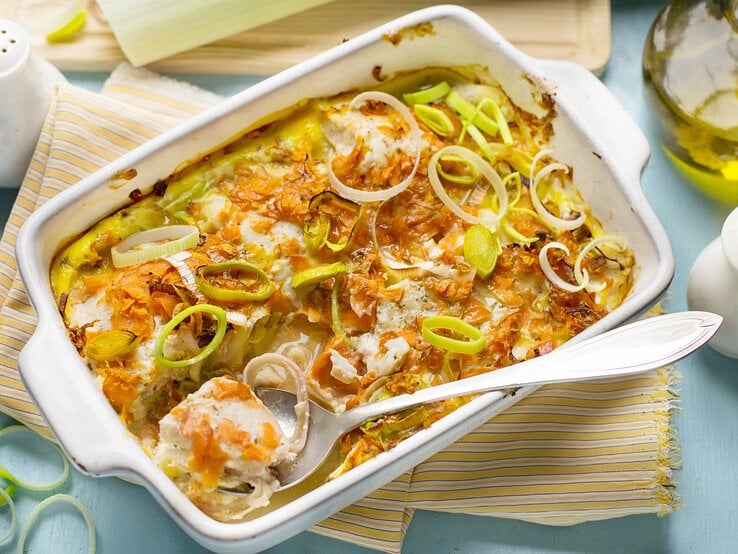 Ofengericht in weißer Auflaufform mit Fisch, Möhren und Lauch. Daneben Salzstreuer, eine Kanne Öl und ein Geschirrtuch.