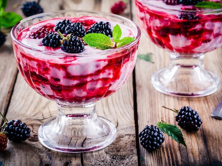 Frontal: Untergrund aus Holz, zwei Dessert-Gläser Brombeer-Vanille-Creme und einige lose Brombeeren sowie ein Dessertlöffel.