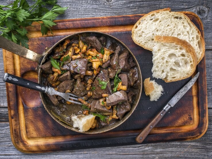 Draufsicht: auf dunklem Untergrund liegt ein Brettchen mit einer rustikalen Pfanne geratene Leber mit Pfifferlingen.