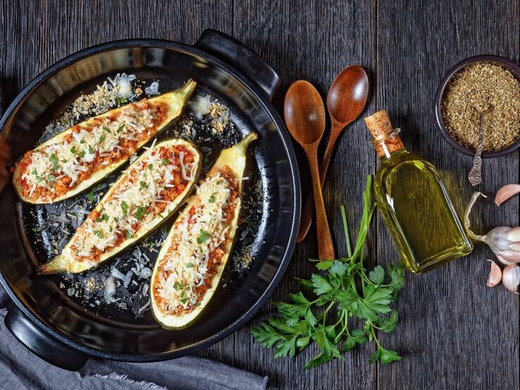 Draufsicht: Gefüllte Zucchini mit Lammhack und Oliven auf einem dunklen Untergrund aus Holz. Daneben liegen zwei Löffel aus Holz, Petersilienzweige, Knoblauchzehen, eine kleine Glasflasche mit Öl und eine schwarze Schale mit einem Gewürz.