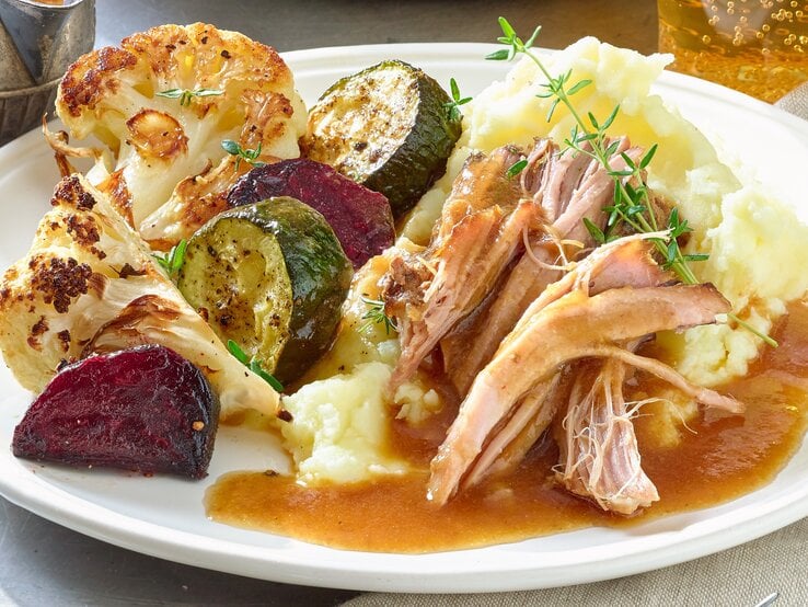 Pulled-Pork-Braten auf einem weißen Teller mit Kartoffelpüree, gebratenem Blumenkohl, Zucchini und roter Bete. Serviert mit Bratensoße. Der Teller steht auf einem Metalltisch neben einem grau-braunen Handtuch.
