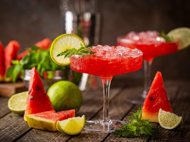 Zwei Gläser Wassermelonen-Daiquiri dekoriert mit Minze und Zitronenscheiben, drumherum frische Minze und Melonenscheiben, dunkler Hintergrund.