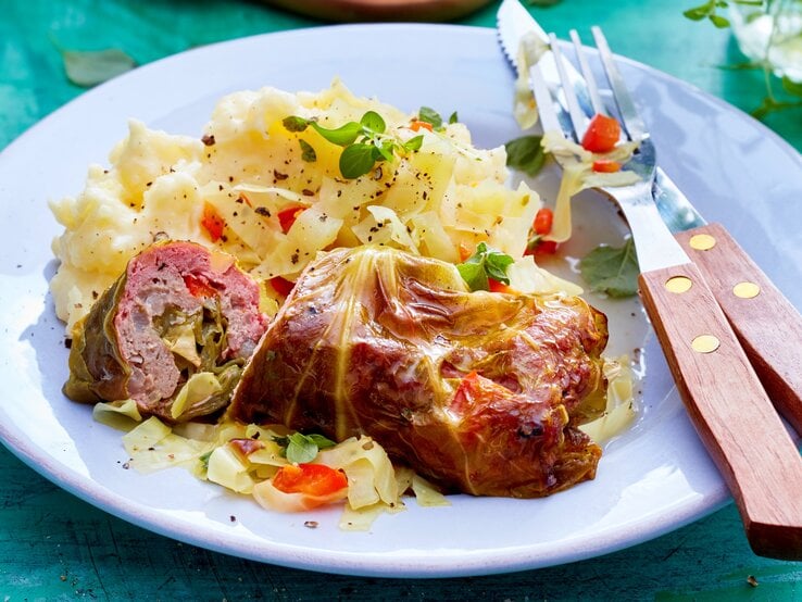 Spitzkohl-Rouladen an Kartoffelpüree auf einem weißen Teller und blauem Untergrund. Alles von schräg oben fotografiert.