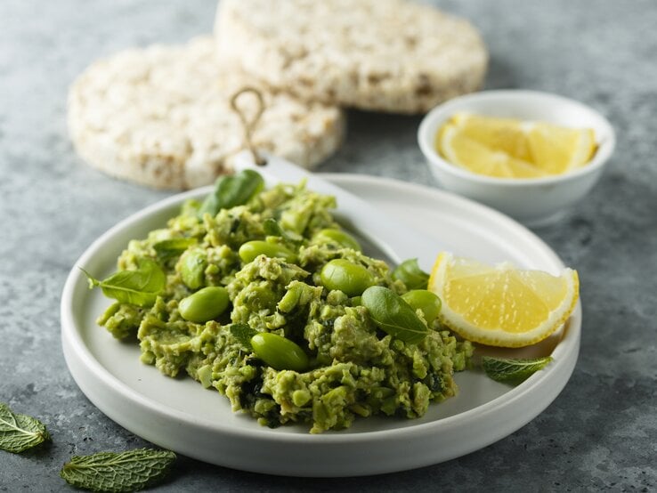 Frontal: eine weiße Schale Edamame-Guacamole steht auf einem grauen Untergrund. Sie ist mit einer Zitronenscheibe garniert. Im Hintergrund liegen Reiswaffeln und noch mehr Zitronenscheiben.