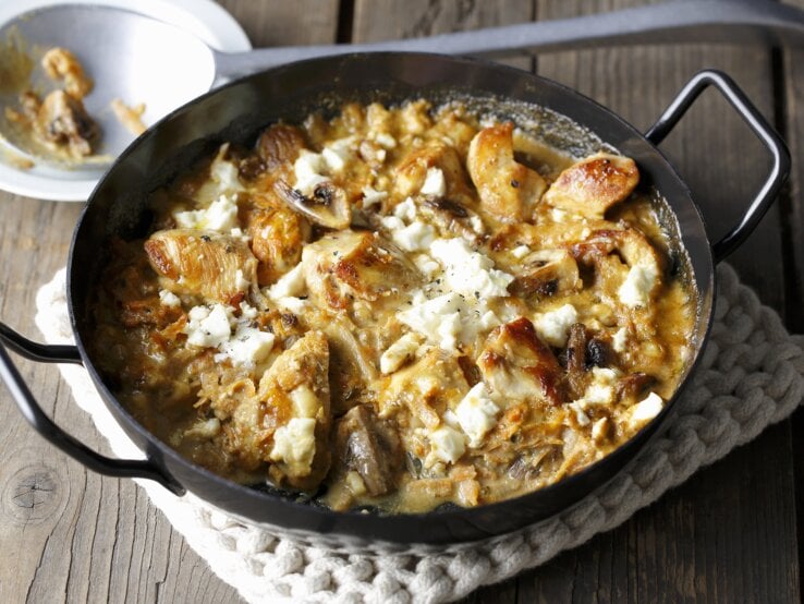 Eine gusseiserne Pfanne mit dem Gericht auf einer beigen Tischdecke und rustikalem Holztisch. Alles schräg oben von der Seite fotografiert.
