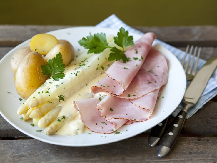 Ein weißer Teller mit Spargel, Kartoffeln, Schinken und Sauce hollandaise auf einem rustikalen Holztisch.