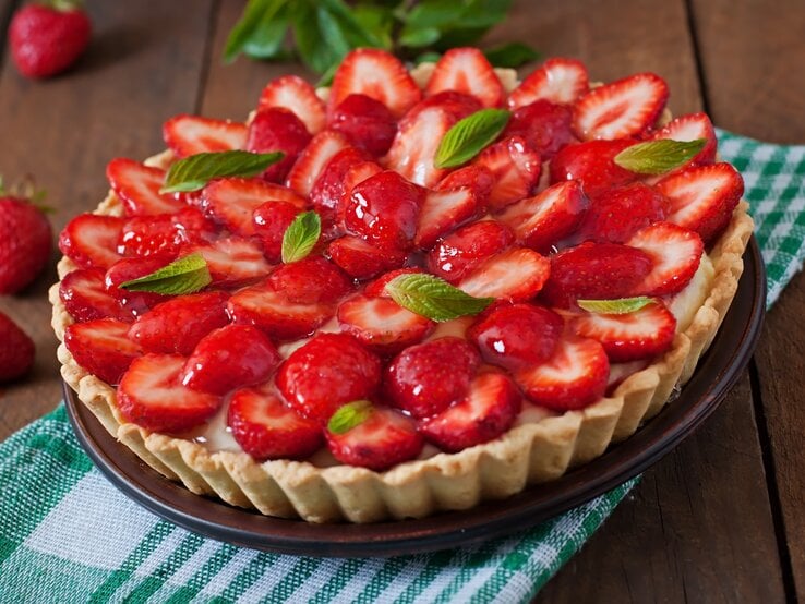 Französische Tarte mit Erdbeeren und Mascarpone-Creme auf einem grün-weiß-kariertem Tuch vor hölzernem Hintergrund, darauf Erdbeeren verteilt.