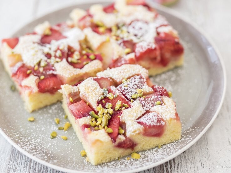 Drei quadratische Stücke Rhabarberkuchen vom Blech auf einem runden Teller.