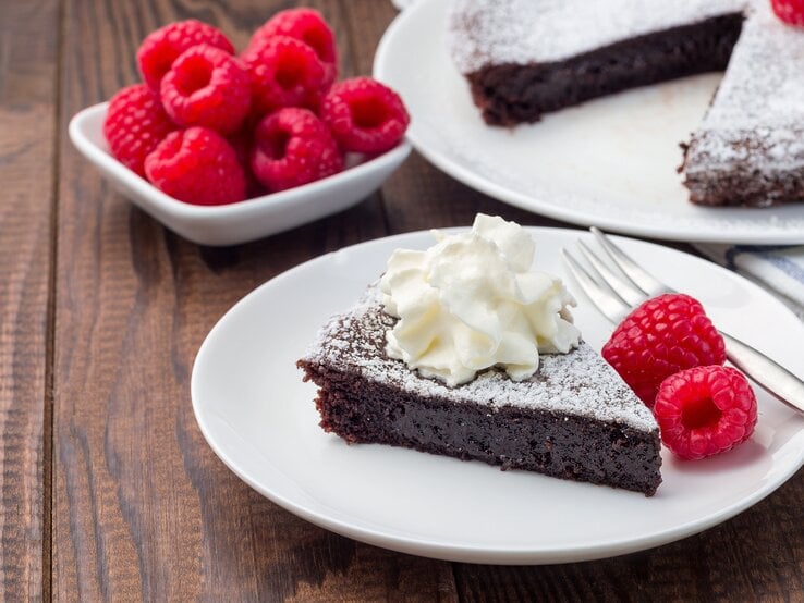 Schokoladenkuchen mit Puderzucker, Sahne und Himbeeren auf weißem Teller in rustikalem Ambiente.  | © Getty Images/iuliia_n
