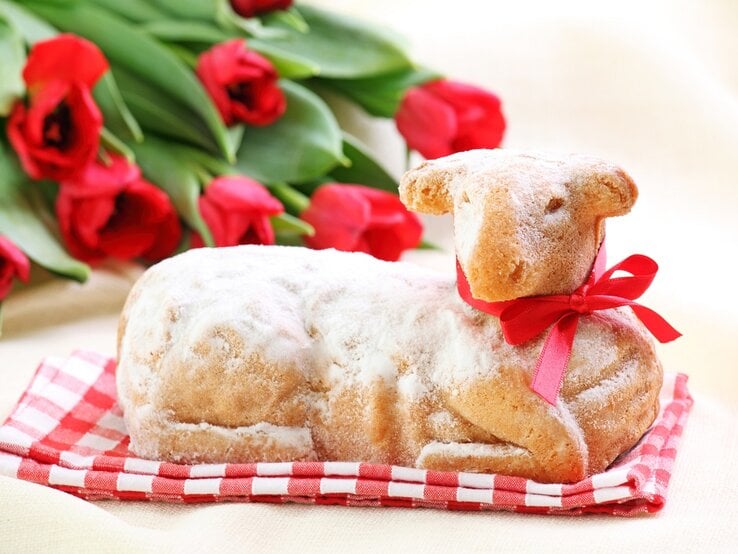 Das Bild zeigt ein Gebäck in Form eines Lamms, das mit Puderzucker bestäubt ist und mit einem roten Band geschmückt ist. Im Hintergrund sind rote Tulpen zu sehen, die auf einem hellen Untergrund liegen. Es sieht nach einem festlichen Essen aus, vielleicht im Zusammenhang mit einem Feiertag wie Ostern. | © istock/egal