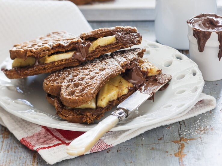 Schoko-Waffeln mit Banane auf einem weißen Teller auf einem Geschirrtuch. Auf dem Teller liegt ein Messer und hinten auf dem Bild ein Glas mit Schoko-Creme. | © StockFood / Young, Andrew