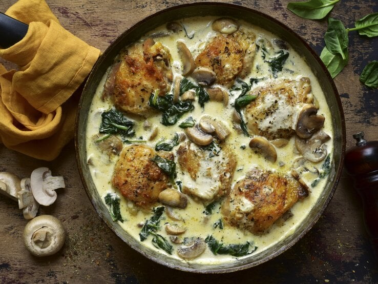 Hähnchenschenkel in Spinat-Pilzrahm-Soße serviert in gusseiserner Pfanne auf hölzernem Untergrund. | © Getty Images/Lilechka75