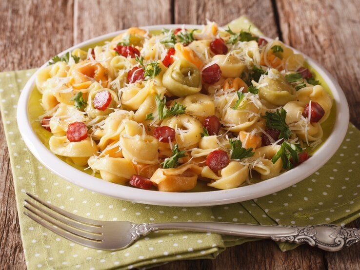 Teller mit Tortellini-Salat mit Salami, Kräutern und Parmesan auf hölzernem Untergrund. 
