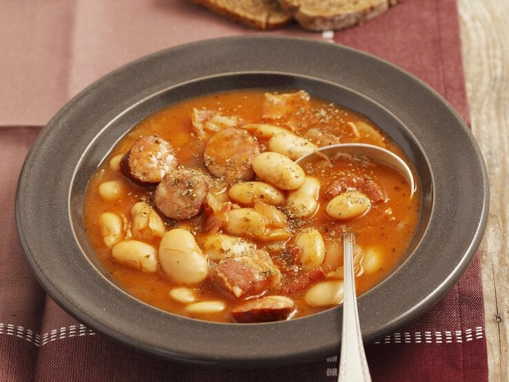 Ein dunkelgrauer Teller mit der Bohnensuppe auf einem rustikalen Holztisch mit roter Tischdecke. | © StockFood/Castilho, Rua