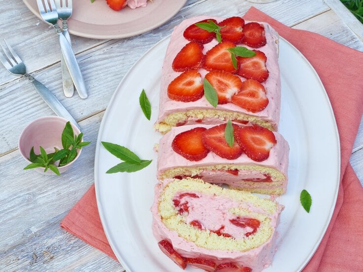Erdbeerrolle mit Buttermilch-Zitronenmousse auf einem weißen Teller auf einem roten Geschirrtuch auf hölzernem Hintergrund. 