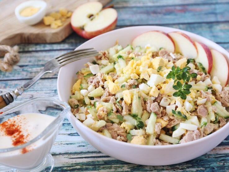 Cremiger Salat aus Ei, Thunfisch und Gurken, mit Apfelscheiben und Kräutern garniert, serviert auf einem Holztisch.