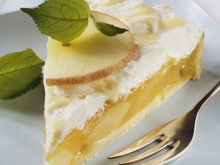 Nahaufnahme eines Stücks Eierlikör-Apfel-Torte mit Vanillecreme, frischen Apfelscheiben und Minzblättern auf einem weißen Teller, daneben eine Kuchengabel