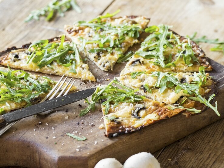 Nahaufnahme vom in Stücke geschnittenen, veganen Flammkuchen mit Rucola und Pilzen.