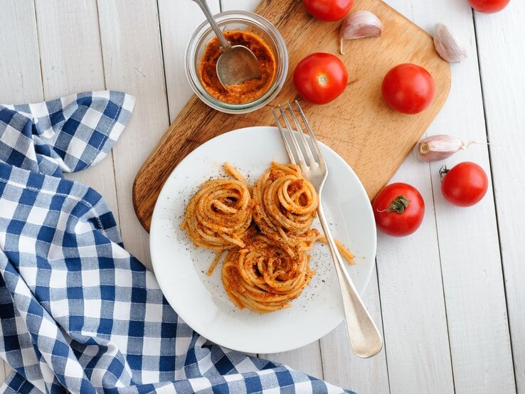 Aufgedrehte Spaghetti-Nester mit Pesto rosso auf weißem Teller. Daneben Tomaten und Knoblauch. 