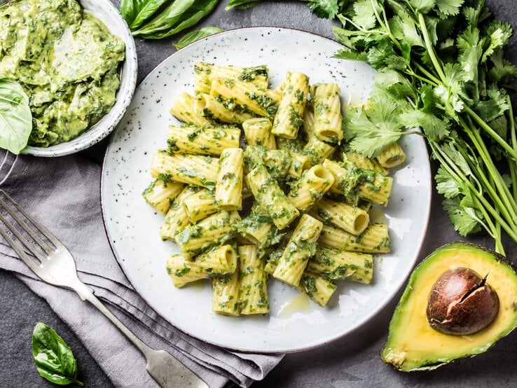 Weißer teller, darauf angerichtet Penne mit einem cremigen Avocado-Pesto. Die Zutaten sind drumherum verteilt. 