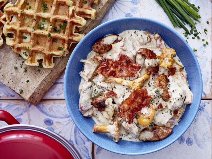 Ein Teller mit Rahmgeschnetzeltem aus Pilzen und Speck, serviert in einer blauen Schale, daneben frische herzhafte Waffeln mit Schnittlauch.
