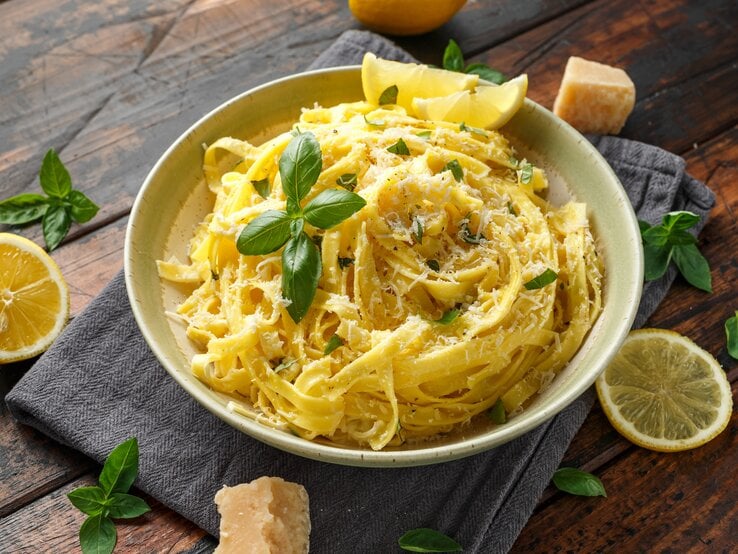 Ein Teller mit Linguine al Limone in einer cremigen Zitronensauce, garniert mit frisch geriebenem Parmesan und Basilikumblättern. Neben dem Teller liegen Zitronenscheiben, frische Basilikumblätter und ein Stück Parmesan auf einem dunklen Holztisch.
