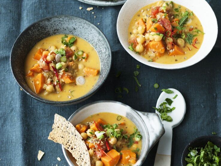 Das Bild zeigt drei Schüsseln, gefüllt mit einer cremigen Suppe, die Stücke von Gemüse und Kichererbsen enthält. Neben den Schüsseln liegen dünne, knusprige Brotscheiben und ein kleiner Teller mit einem Löffel. Im Vordergrund steht eine kleine Schale mit frisch geschnittenen Kräutern. Die Szene vermittelt den Eindruck einer liebevollen und vielseitigen Mahlzeit.