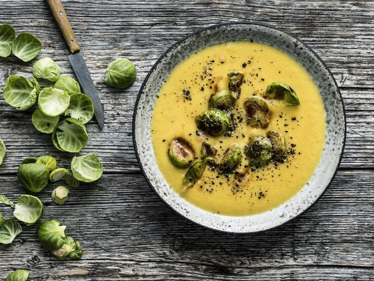 Draufsicht: Hölzerner Untergrund auf der ein grauer Steingutteller mit Steckrübensuppe und Rosenkohl-Topping steht. Links daneben noch Schalen frischer Rosenkohlröschen und ein scharfes Messer mit Holzgriff. | © StockFood / Gräfe & Unzer Verlag / Schardt, Wolfgang