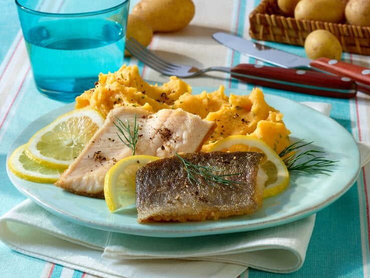 Ein Teller Kartoffel-Kürbis-Püree zu Forellenfilet auf einem Tisch / Besteck / Glas / Tuck / rohe Kartoffeln im Korb