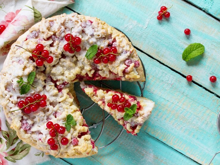 Johannisbeerkuchen mit Streusel auf einem Kuchengitter vor blauem Hintergrund. Links eine Tischdecke. | © iStock/as117pxz